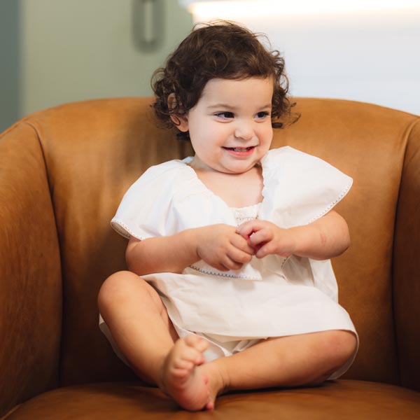 Smiling toddler in a white dress sitting on a brown chair in Dripping Springs, TX